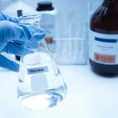 Ręka w niebieskiej rękawiczce trzyma kolbę z płynem, na której widnieje napis "Ethyl ether". W tle ciemna butelka z substancją chemiczną.
