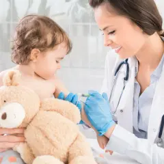 Uśmiechnięta lekarka podaje szczepionkę MMR małemu dziecku, które przytula pluszowego misia.