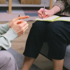Dwie osoby siedzą naprzeciwko siebie. Jedna z nich, w jasnej bluzie, gestykuluje, druga, w kratkę, notuje coś w zeszycie. To może być sesja terapeutyczna lub kurs terapeuty zajęciowego.