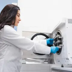 Technik sterylizacji medycznej zarobki: kobieta w białym fartuchu i niebieskich rękawiczkach wkłada tacki do autoklawu.