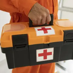 Pomarańczowa skrzynka z czerwonym krzyżem, trzymana przez osobę w pomarańczowym kombinezonie.