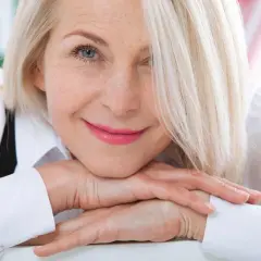 Uśmiechnięta kobieta z blond włosami, opierająca podbródek na dłoniach. Jej spojrzenie jest ciepłe, a makijaż podkreśla jej naturalne piękno. Veoza.