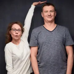Kobieta z wyrazem zaskoczenia na twarzy próbuje zmierzyć wzrost mężczyzny, który wydaje się mieć gigantyzm.