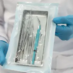 Ręce w niebieskich rękawiczkach trzymają opakowanie ze sterylnymi narzędziami medycznymi.