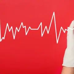 Biała linia EKG na czerwonym tle, przechodząca przez czerwone serce trzymane w białych rękawiczkach. Symbolizuje troskę o serce i jego rytm, ostrzegając przed migotaniem komór.