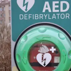 Zielony AED (defibrylator) z symbolem serca i pioruna na tle miejskim.
