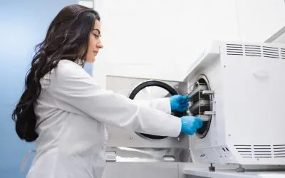 Technik sterylizacji medycznej zarobki: kobieta w białym fartuchu i niebieskich rękawiczkach wkłada tacki do autoklawu.