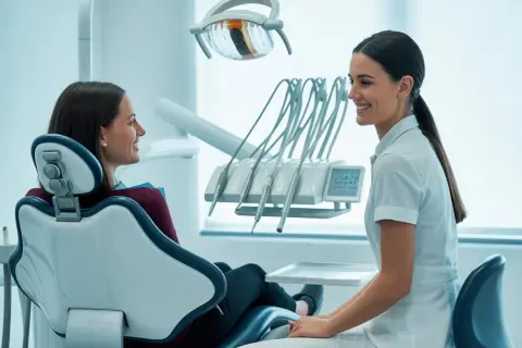 Uśmiechnięta higienistka stomatologiczna rozmawia z pacjentką siedzącą na fotelu dentystycznym.