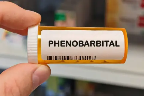 Dłoń trzyma buteleczkę z lekiem. Na etykiecie widnieje napis "PHENOBARBITAL" i kod kreskowy.