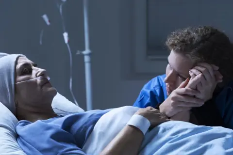 Chora kobieta w łóżku szpitalnym, podłączona do aparatury, trzyma za rękę bliską osobę. Czy leczenie paliatywne to koniec?