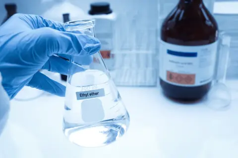 Ręka w niebieskiej rękawiczce trzyma kolbę z płynem, na której widnieje napis "Ethyl ether". W tle ciemna butelka z substancją chemiczną.