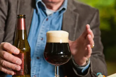 Mężczyzna trzyma butelkę piwa obok pełnego kufla. Czy po szczepieniu można pić alkohol? To pytanie nurtuje wielu.
