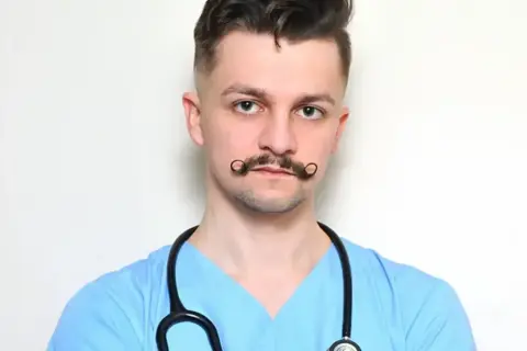 Młody rezydent w niebieskiej bluzie medycznej, ze stetoskopem na szyi i charakterystycznymi wąsami.