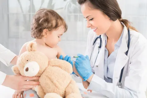 Uśmiechnięta lekarka podaje szczepionkę MMR małemu dziecku, które przytula pluszowego misia.