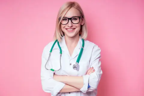 Uśmiechnięta pielęgniarka w okularach i białym fartuchu, z zielonym stetoskopem na szyi, stoi ze skrzyżowanymi rękami.