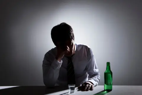 Zmartwiony mężczyzna w białej koszuli i krawacie, z ręką na czole, siedzi przy stole z butelką zielonego alkoholu i kieliszkiem. Zastanawia się, ile trwa zatrucie alkoholowe.