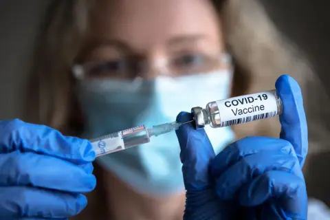 Osoba w niebieskich rękawiczkach przygotowuje szczepionki na covid, pobierając płyn do strzykawki z fiolki z napisem "COVID-19 Vaccine".