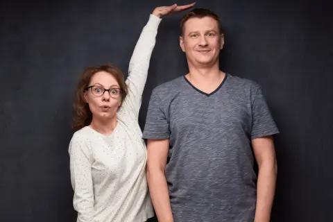 Kobieta z wyrazem zaskoczenia na twarzy próbuje zmierzyć wzrost mężczyzny, który wydaje się mieć gigantyzm.