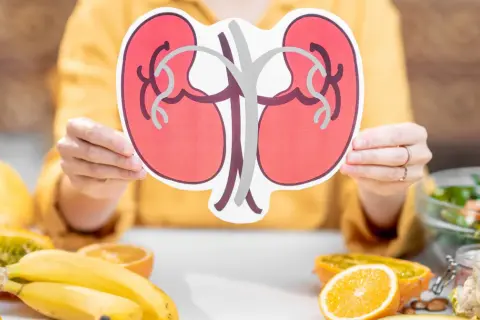 Osoba trzyma symbol nerek, a na stole leżą owoce i warzywa. Zastanawiasz się, co pić na chore nerki? Woda i zdrowe napoje to podstawa.