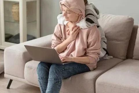 Kobieta z dzieckiem na kolanach pracuje przy laptopie. Dziecko ją przytula, szukając wsparcia, jak u psychoonkologa.