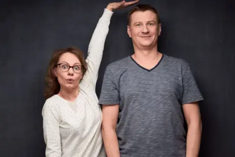 Kobieta z wyrazem zaskoczenia na twarzy, z ręką uniesioną do góry, obok wysokiego mężczyzny. Może to być wizualizacja różnic wzrostu, czasem kojarzonych z zespołem Marfana.