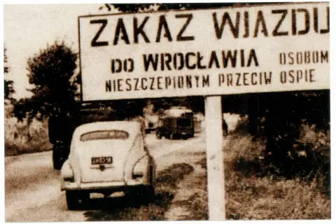Znak "Zakaz wjazdu do Wrocławia osobom nieszczepionym przeciw ospie". W tle samochód i drzewa.