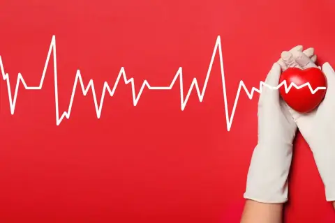 Biała linia EKG na czerwonym tle, przechodząca przez czerwone serce trzymane w białych rękawiczkach. Symbolizuje troskę o serce i jego rytm, ostrzegając przed migotaniem komór.