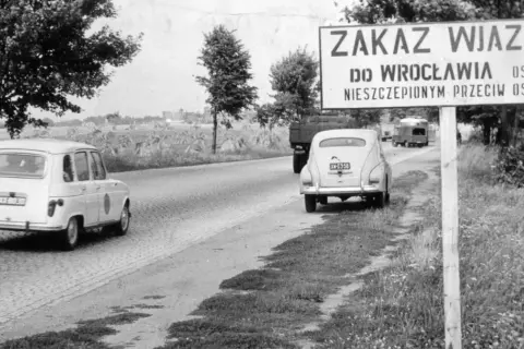 Znak "Zakaz wjazdu do Wrocławia osobom nieszczepionym przeciw ospie" przy drodze. W tle samochody i pola.
