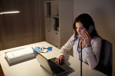 Młoda kobieta, pracująca jako dyspozytor, odbiera telefon przy biurku z laptopem i dokumentami.