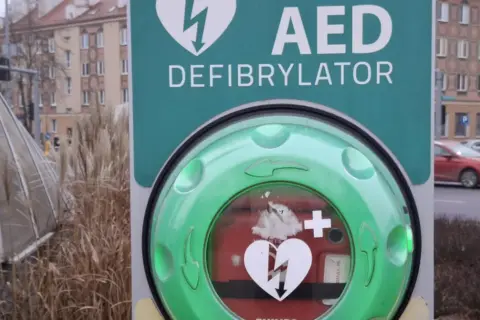 Zielony AED (defibrylator) z symbolem serca i pioruna na tle miejskim.