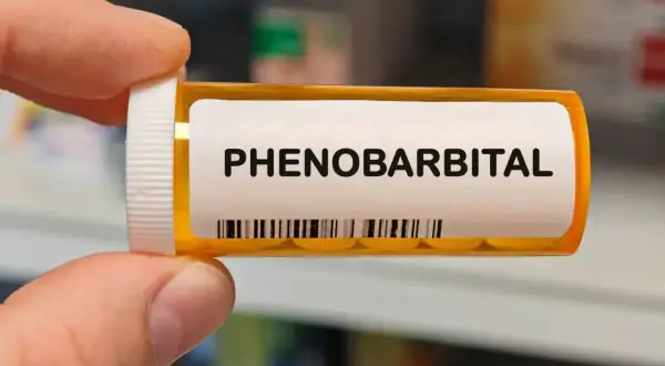 Dłoń trzyma buteleczkę z lekiem. Na etykiecie widnieje napis "PHENOBARBITAL" i kod kreskowy.