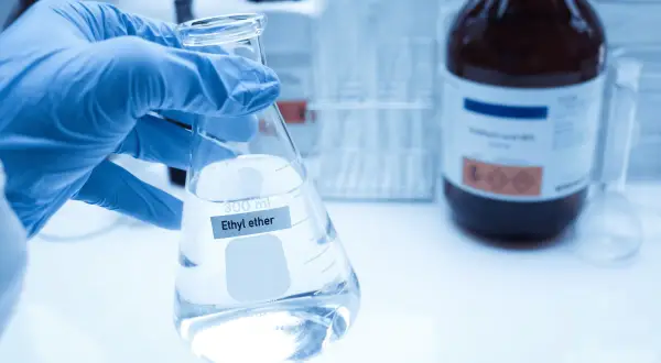 Ręka w niebieskiej rękawiczce trzyma kolbę z płynem, na której widnieje napis "Ethyl ether". W tle ciemna butelka z substancją chemiczną.