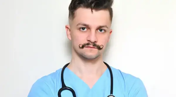 Młody rezydent w niebieskiej bluzie medycznej, ze stetoskopem na szyi i charakterystycznymi wąsami.