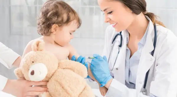 Uśmiechnięta lekarka podaje szczepionkę MMR małemu dziecku, które przytula pluszowego misia.