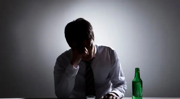 Zmartwiony mężczyzna w białej koszuli i krawacie, z ręką na czole, siedzi przy stole z butelką zielonego alkoholu i kieliszkiem. Zastanawia się, ile trwa zatrucie alkoholowe.