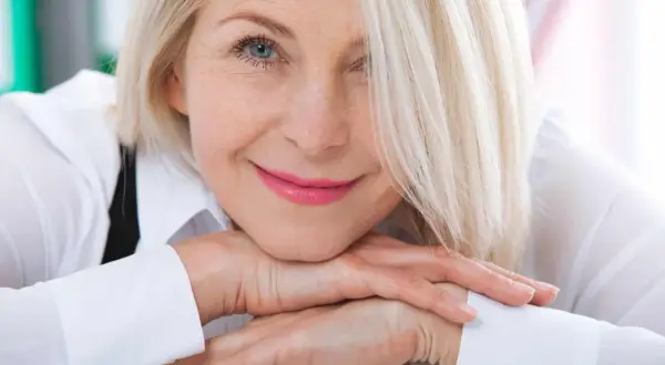 Uśmiechnięta kobieta z blond włosami, opierająca podbródek na dłoniach. Jej spojrzenie jest ciepłe, a makijaż podkreśla jej naturalne piękno. Veoza.