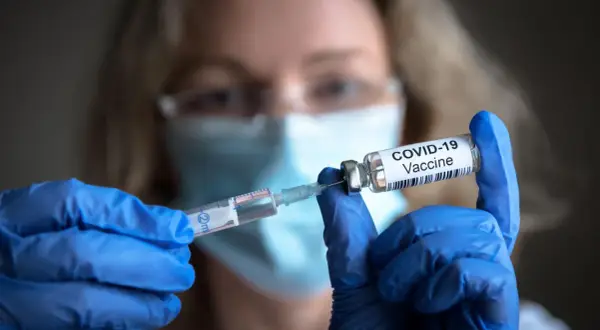 Osoba w niebieskich rękawiczkach przygotowuje szczepionki na covid, pobierając płyn do strzykawki z fiolki z napisem "COVID-19 Vaccine".