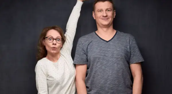 Kobieta z wyrazem zaskoczenia na twarzy, z ręką uniesioną do góry, obok wysokiego mężczyzny. Może to być wizualizacja różnic wzrostu, czasem kojarzonych z zespołem Marfana.