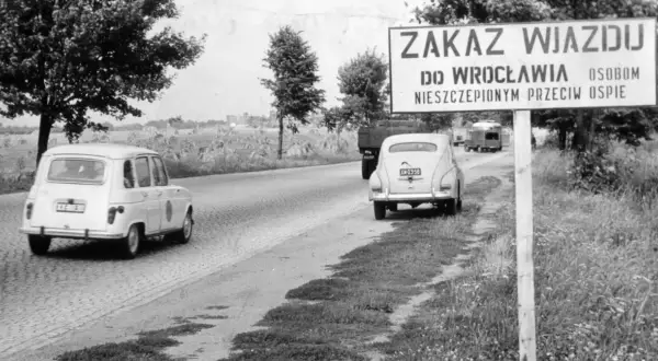Znak "Zakaz wjazdu do Wrocławia osobom nieszczepionym przeciw ospie" przy drodze. W tle samochody i pola.