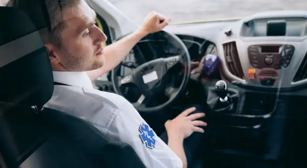 Kierowca karetki w białej koszuli z symbolem ratunkowym, za kierownicą. Zastanawiasz się, ile zarabia kierowca karetki?