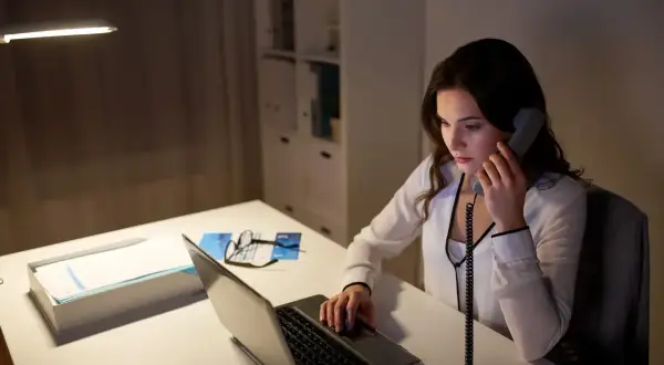 Młoda kobieta, pracująca jako dyspozytor, odbiera telefon przy biurku z laptopem i dokumentami.