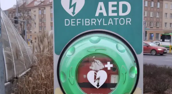 Zielony AED (defibrylator) z symbolem serca i pioruna na tle miejskim.