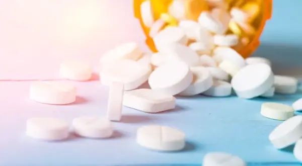 Białe tabletki, w tym leki opioidowe, rozsypane z pomarańczowego pojemnika na niebieskim tle.