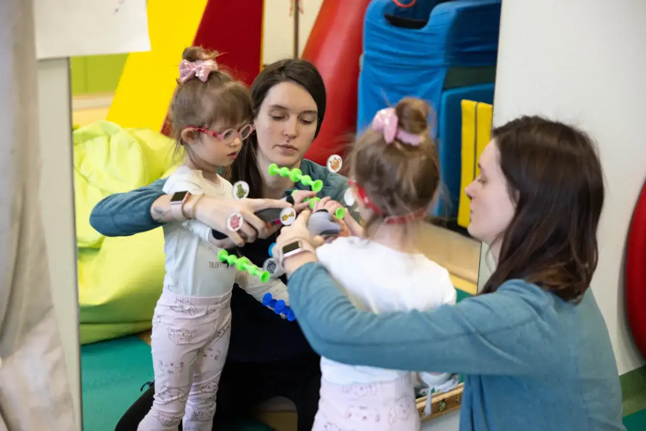 Dwie kobiety pomagają dziewczynce z artrogrypozą w ćwiczeniach z kolorowymi klockami.