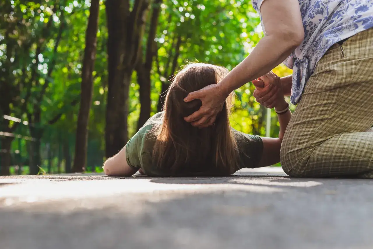 Osoba leży na ziemi, druga osoba próbuje jej pomóc. Może to być atak epilepsji.