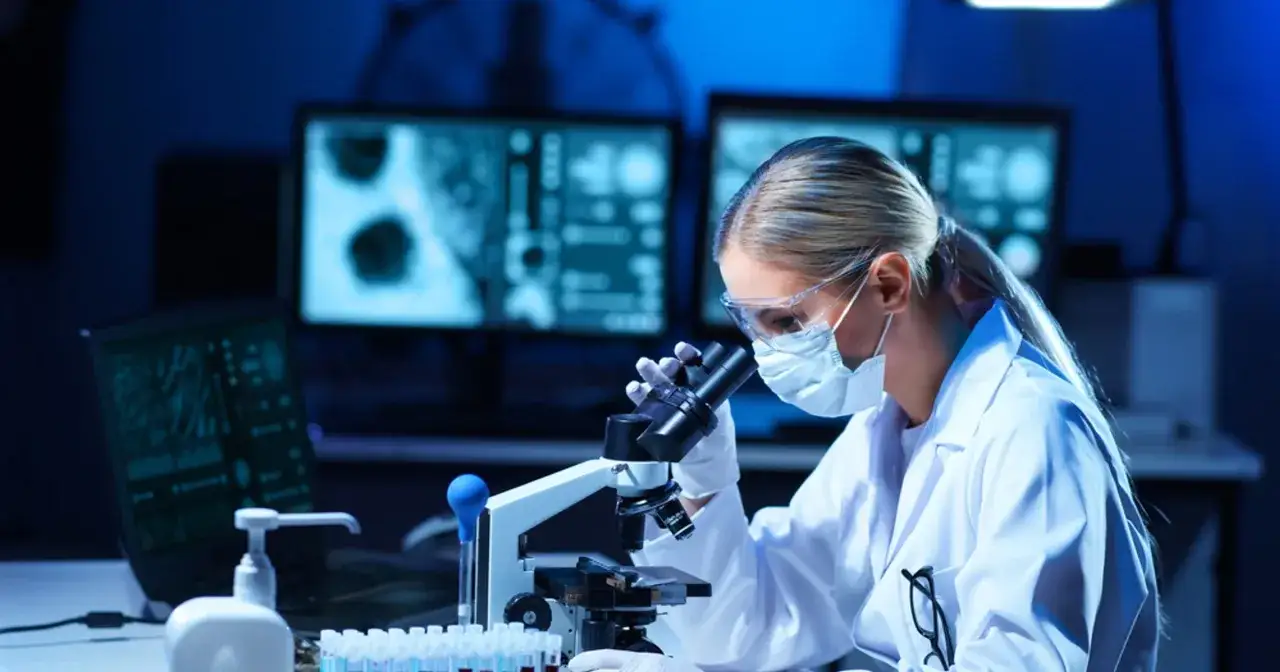 Diagnosta laboratoryjny zarobki: młoda kobieta w okularach i masce pracuje przy mikroskopie w ciemnym laboratorium, analizując próbki.