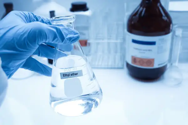 Ręka w niebieskiej rękawiczce trzyma kolbę z płynem, na której widnieje napis "Ethyl ether". W tle ciemna butelka z substancją chemiczną.